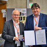 Zwei Männer, einer mit einer Medaille, der andere mit einer Urkunde, stehen in einem Hörsaal.