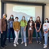 Studierende tragen Masken und stehen vor einem Projektorbildschirm 